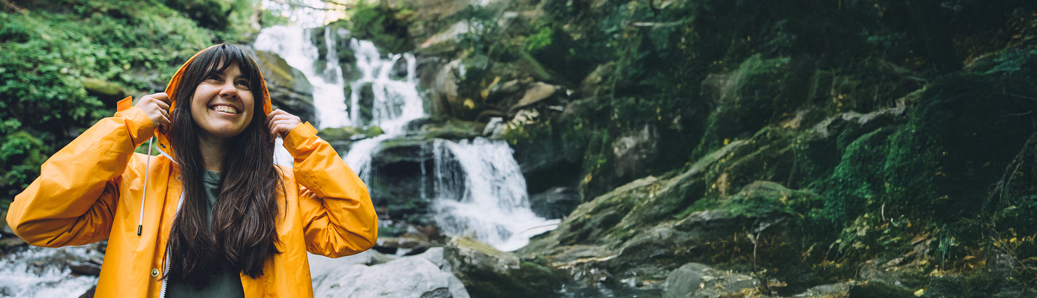 Eine lächelnde Frau in einer gelben Regenjacke steht in der Nähe eines felsigen Wasserfalls, umgeben von üppig grünem Moos und Bäumen, und genießt die freie Natur.