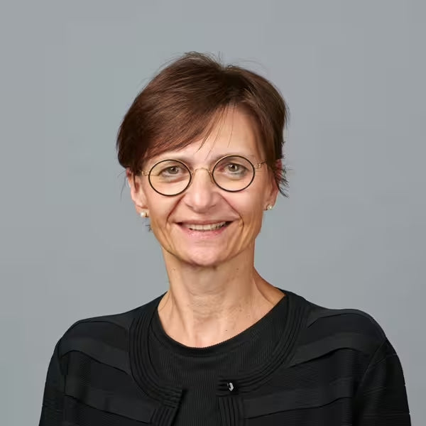 Eine Frau mit kurzen braunen Haaren, einer runden Brille und einem schwarzen Oberteil lächelt vor einem schlichten grauen Hintergrund in die Kamera.