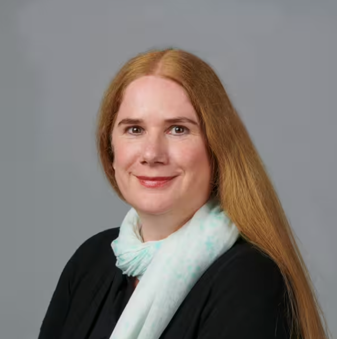 Eine Frau mit langem, glattem, rotem Haar, einem schwarzen Oberteil und einem hellen Schal trägt, lächelt vor einem schlichten grauen Hintergrund in die Kamera.