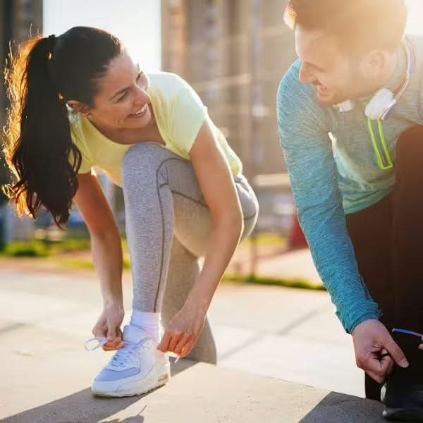 Eine Frau und ein Mann in Sportkleidung lächeln sich an, während sie draußen im Sonnenlicht ihre Schnürsenkel binden und sehen voller Energie aus – vielleicht dank gse.organic.supplements Vino C® - OPC (Bio).