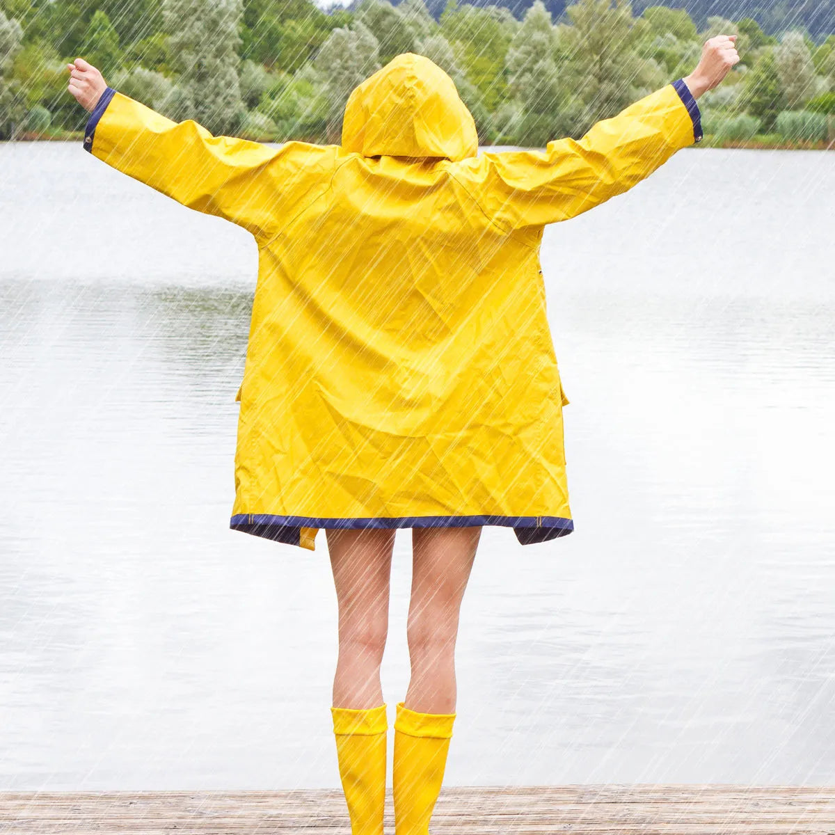Eine Person in gelbem Regenmantel und Gummistiefeln steht mit ausgestreckten Armen auf einem Holzsteg und blickt auf einen im Regen liegenden See. Im Hintergrund sind üppige Bäume und Hügel zu sehen.