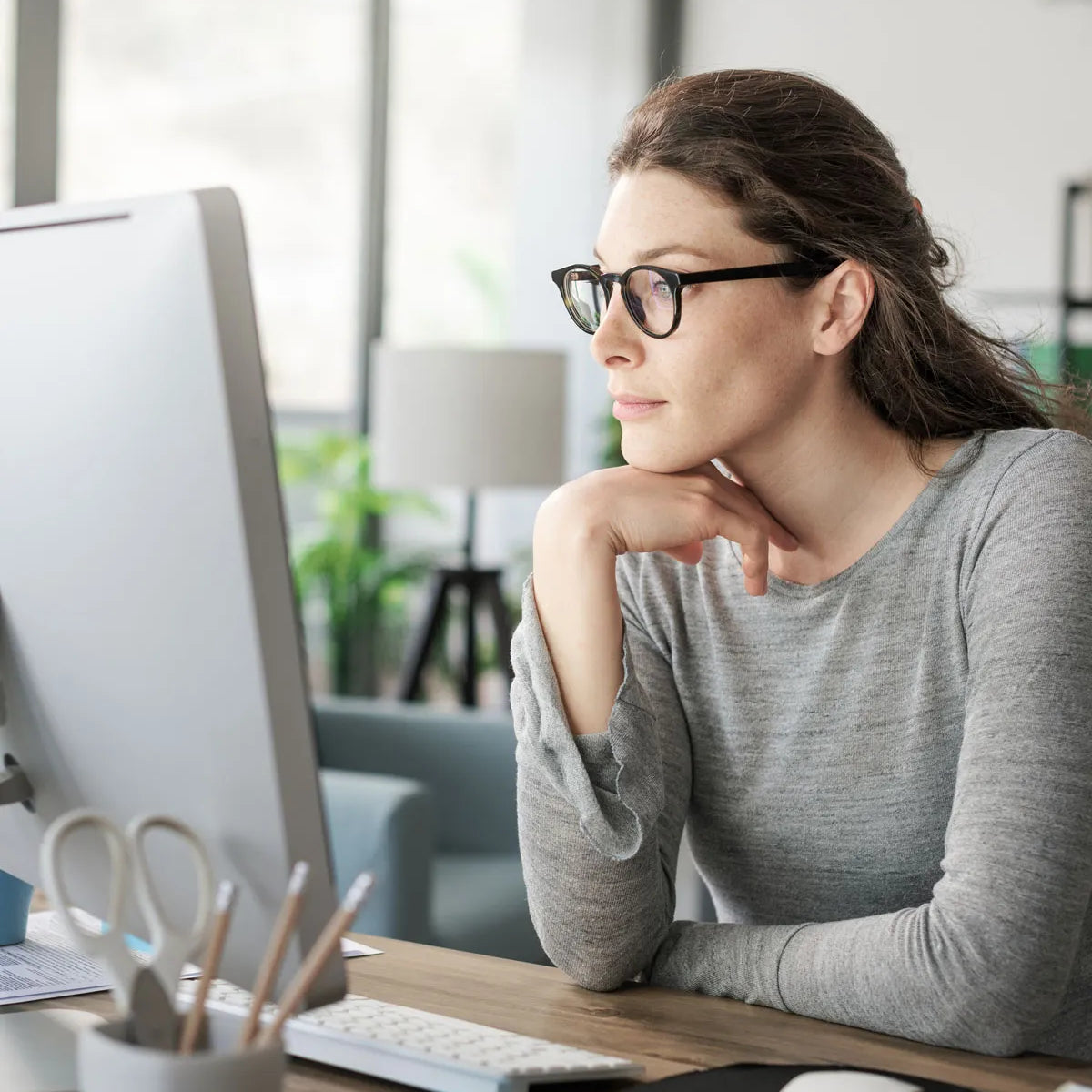 Eine Frau mit Brille und grauem Hemd sitzt an einem Schreibtisch und blickt nachdenklich auf einen Computermonitor. Büromaterial und eine Tastatur liegen auf dem Schreibtisch. Im Hintergrund ist ein helles, modernes Büro zu sehen.