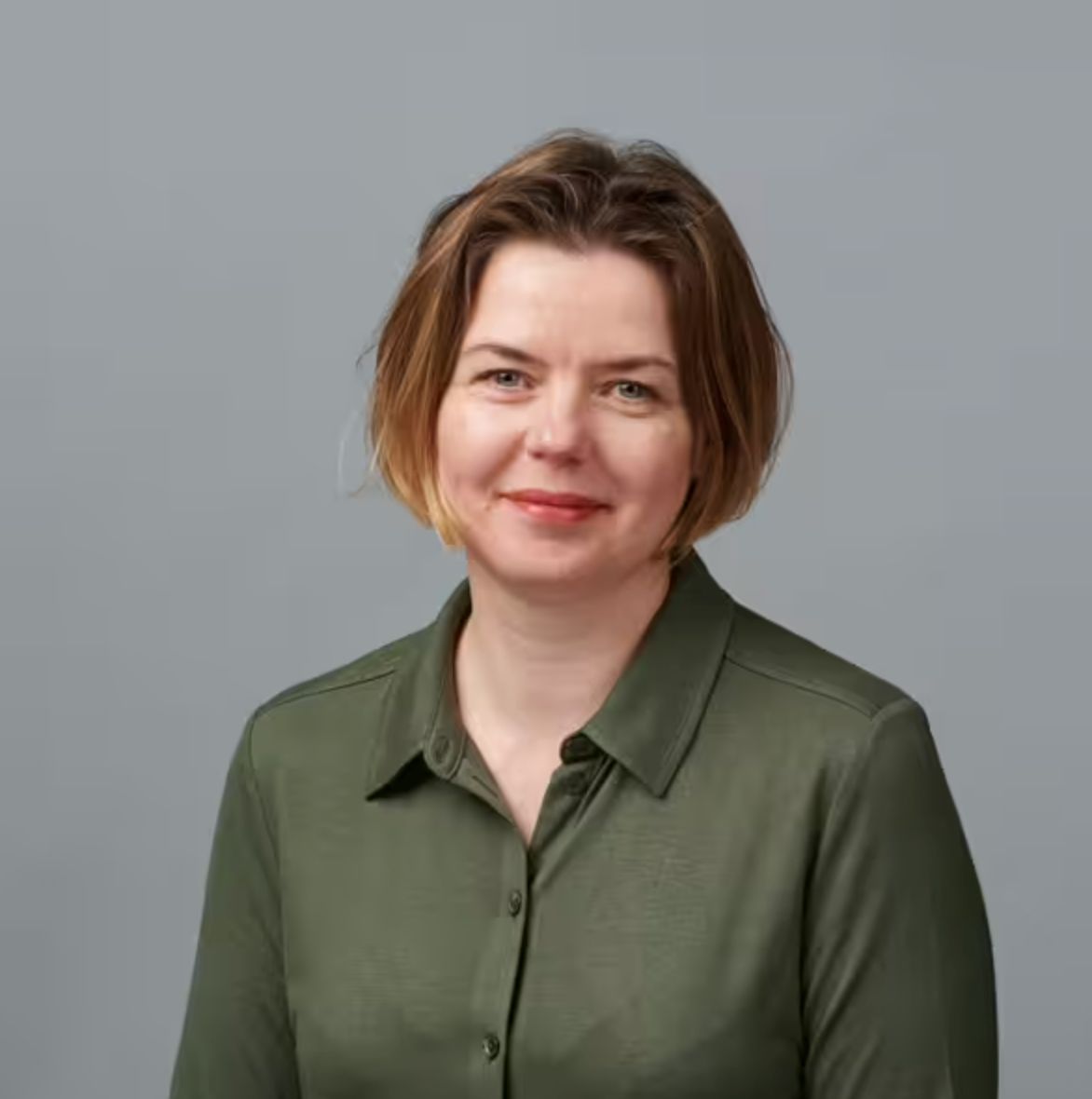 Eine Frau mit kurzen, hellbraunen Haaren und heller Haut lächelt sanft in die Kamera. Sie trägt ein zugeknöpftes olivgrünes Hemd und posiert vor einem schlichten, hellgrauen Hintergrund.