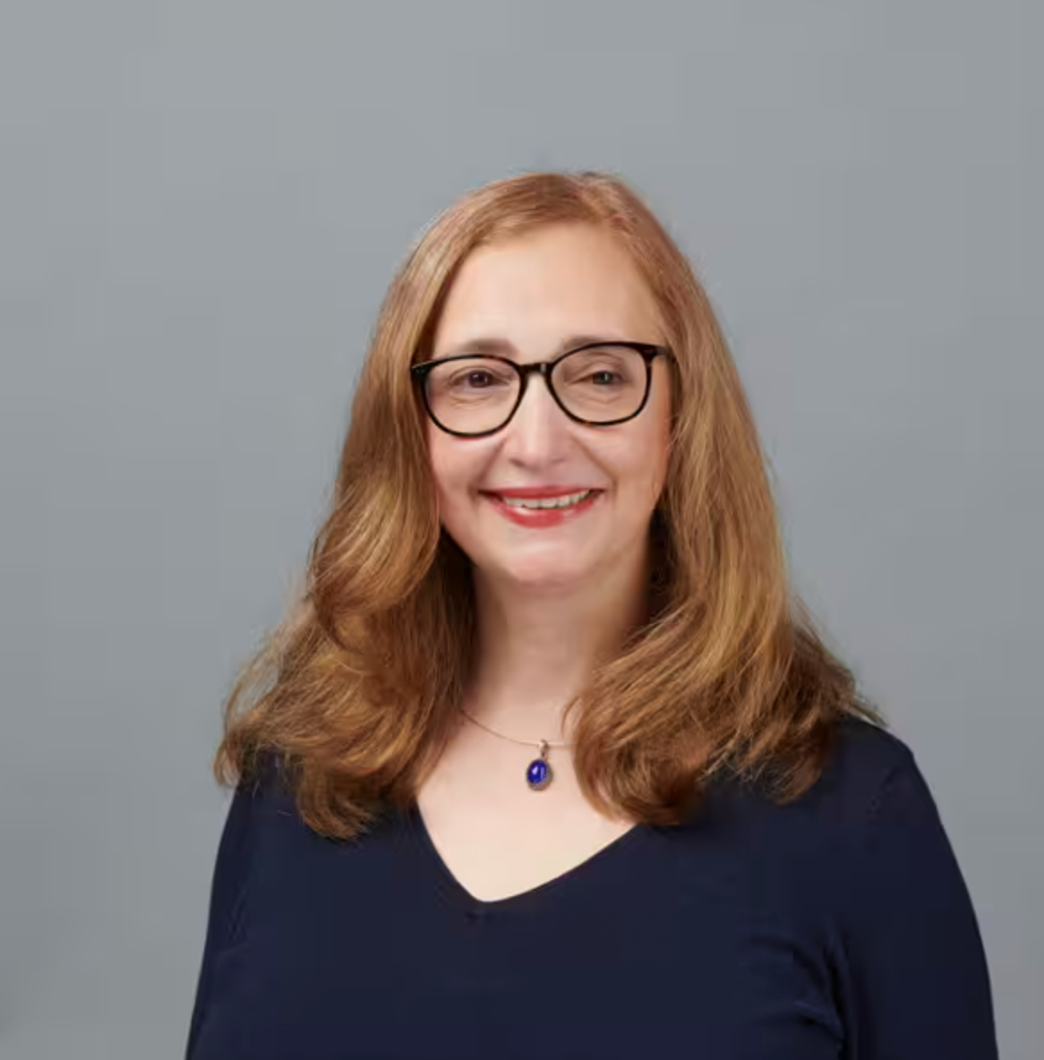 Eine Frau mit schulterlangem hellbraunem Haar, einer Brille, einem marineblauen Oberteil und einer Halskette mit einem blauen Anhänger, lächelt vor einem schlichten grauen Hintergrund.